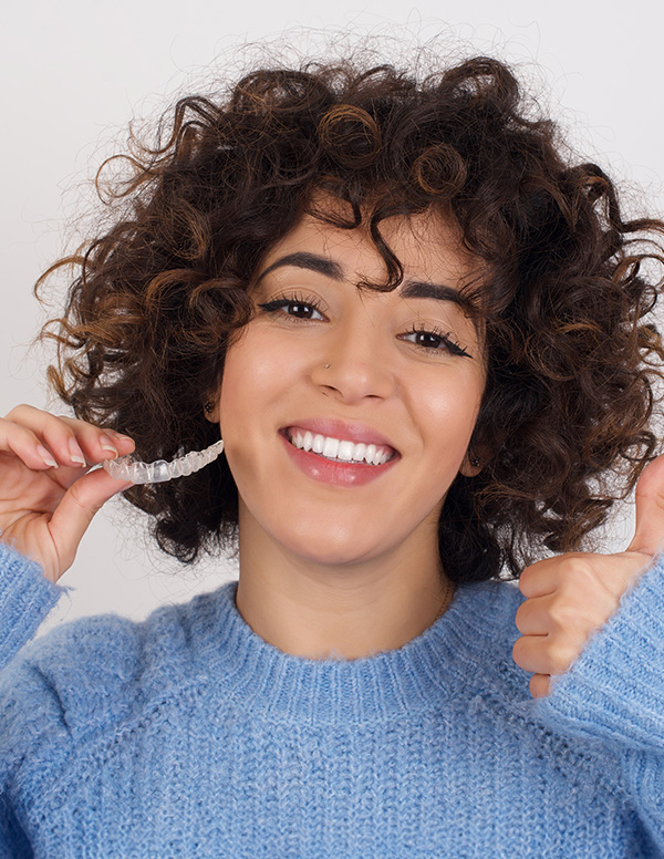A woman with short curly hair, smiling broadly, holding a toothbrush in her right hand.
