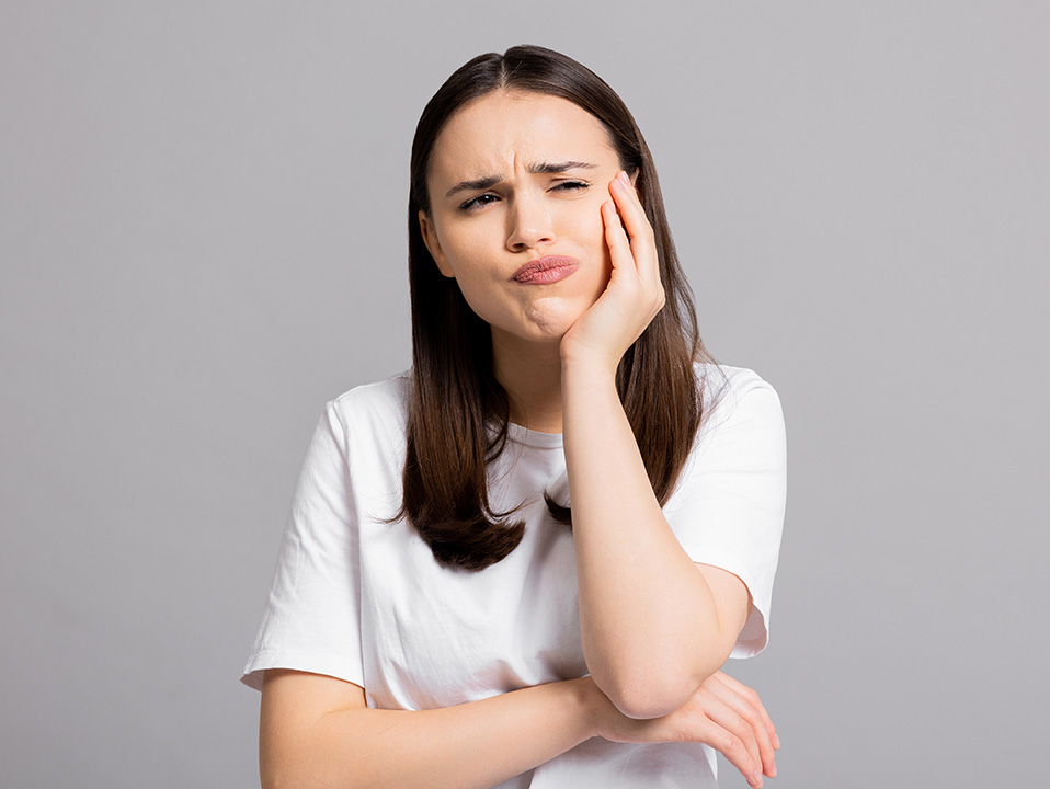 Woman with hand on chin, making a funny face, wearing white t-shirt and dark pants.